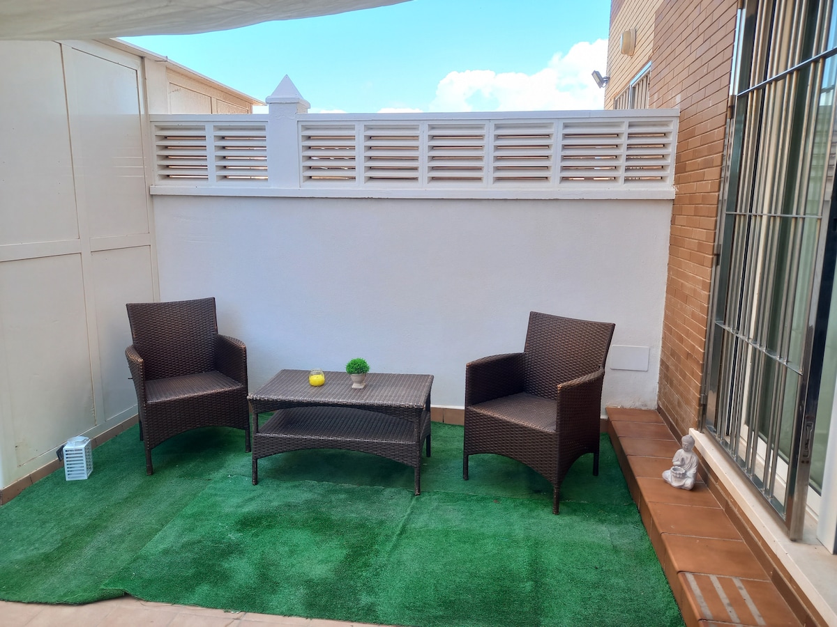 A small patio area is depicted, featuring two wicker chairs and a low table set on a grassy surface. A low white wall provides privacy, while a portion of the adjacent building is visible. Soft lighting and clear skies create a bright environment.