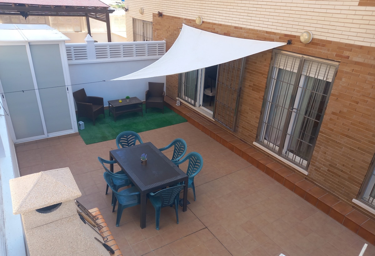 A spacious terrace features a large dining table surrounded by teal chairs, providing ample seating. Two cozy lounge chairs are set on the green turf area. A sunshade offers protection from sunlight, while sliding doors lead into the interior living spaces.