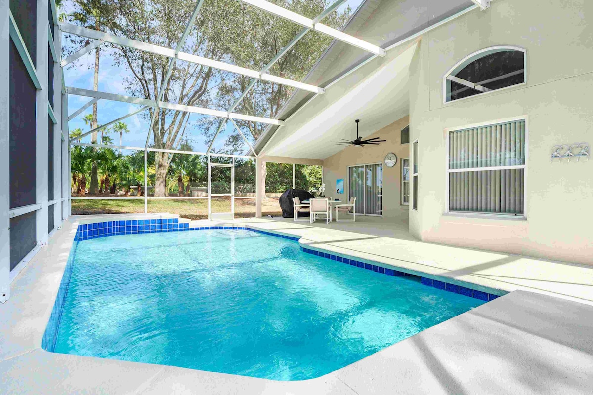 The spacious enclosed lanai features a private pool with crystal clear water, offering a relaxing retreat. Sunlight filters through the screen enclosure, illuminating the area. Comfortable seating is available nearby, surrounded by greenery, enhancing the serene atmosphere of the space.