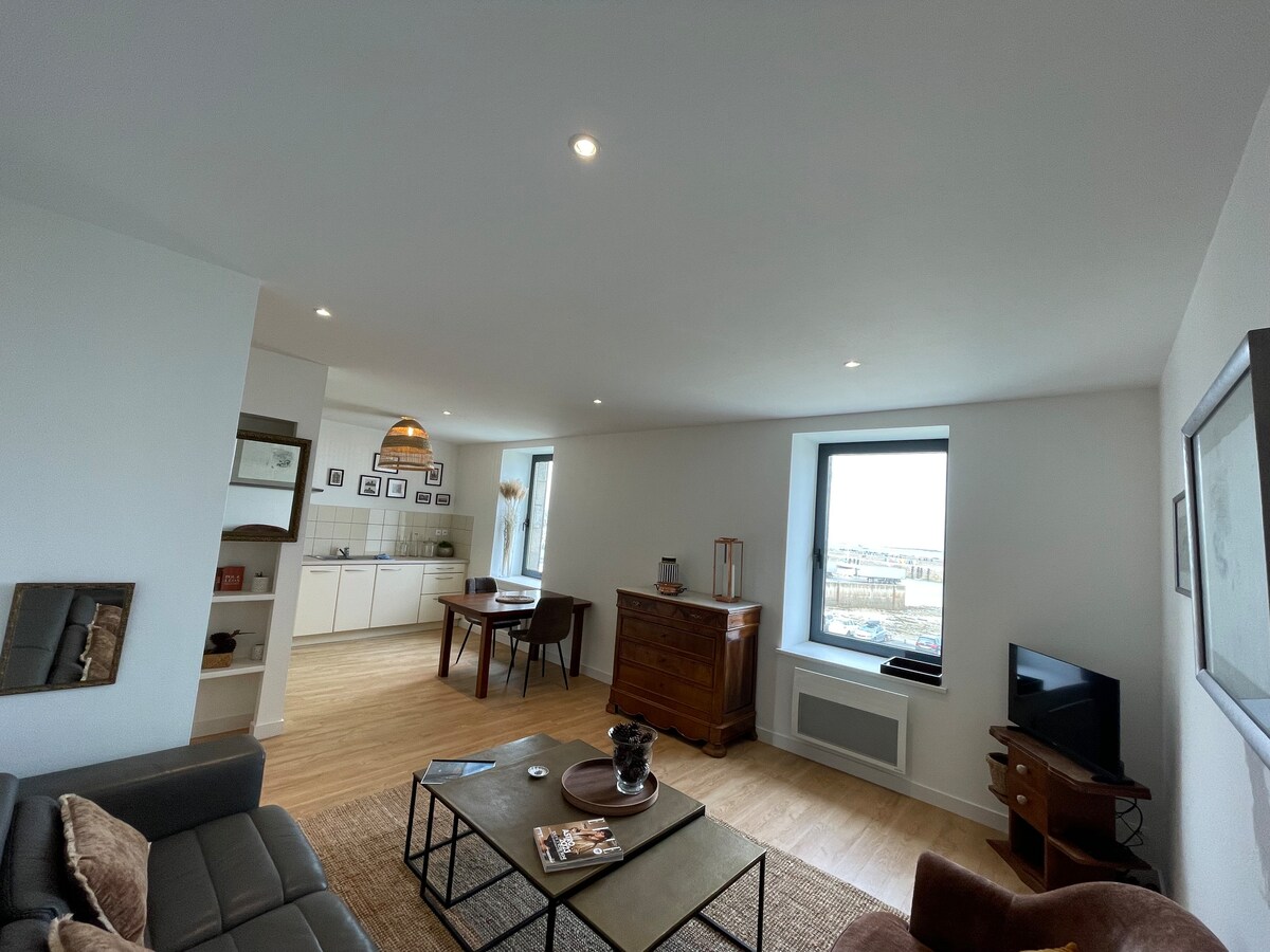 The spacious living area features a comfortable sofa and an armchair, with a wooden coffee table and a television for entertainment. A dining table is positioned nearby with seating for four. Natural light illuminates the room through a large window, showcasing views outside.