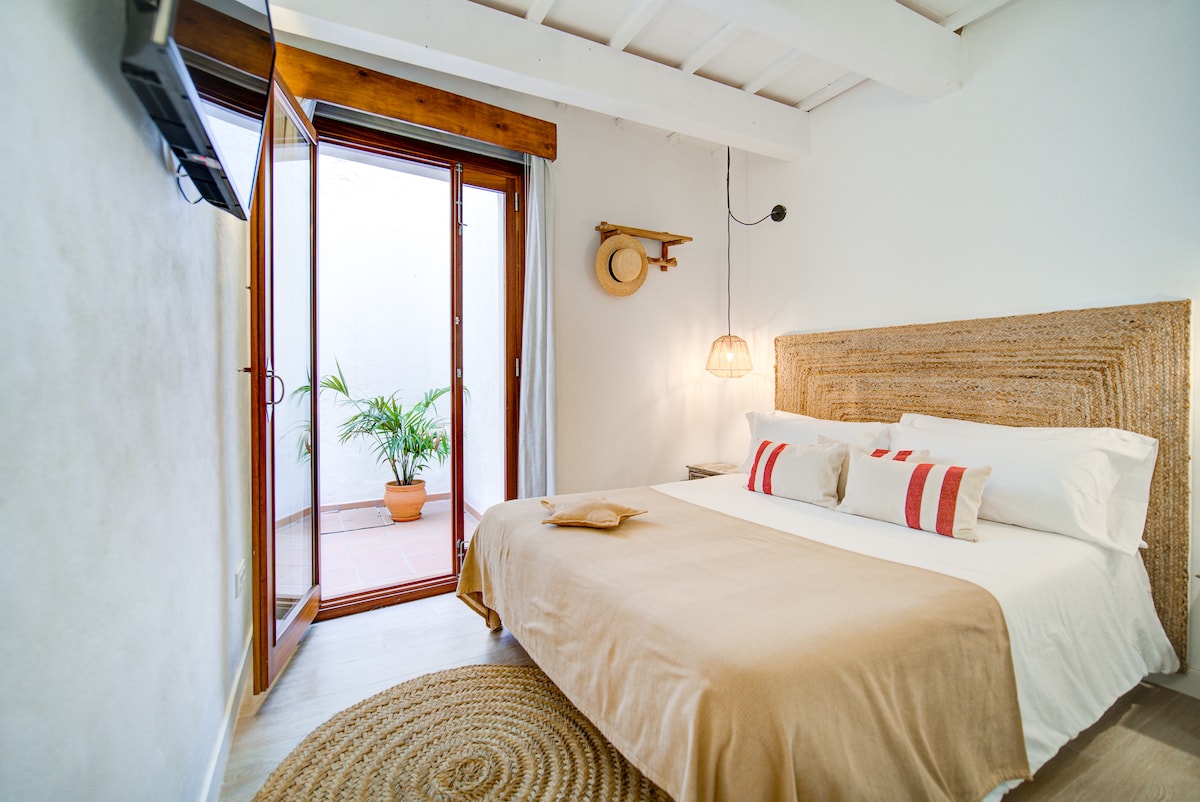 A spacious bedroom features a large bed with neutral linens and decorative pillows. Natural light enters through double doors opening to a charming patio, where a potted plant adds a touch of greenery. A woven wall hanging and pendant light provide subtle decor.