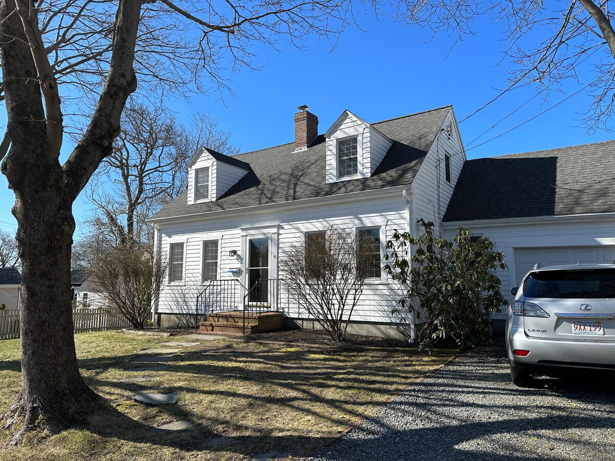 A charming two-story house is featured, with white siding and a dark roof. A welcoming front porch is visible, flanked by several windows. A well-maintained lawn leads to a gravel driveway with a parked vehicle, indicating convenient access.