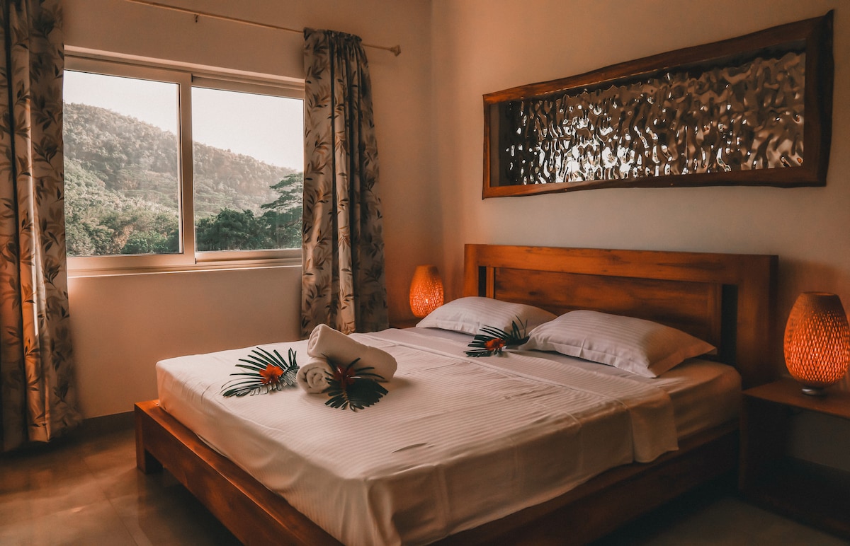 A bright sleeping area is furnished with a spacious bed, topped with neatly arranged linens and decorative towels. Two bedside lamps provide soft illumination, while a large window allows natural light to enter, offering views of the surrounding greenery. Textured wall decor adds interest.