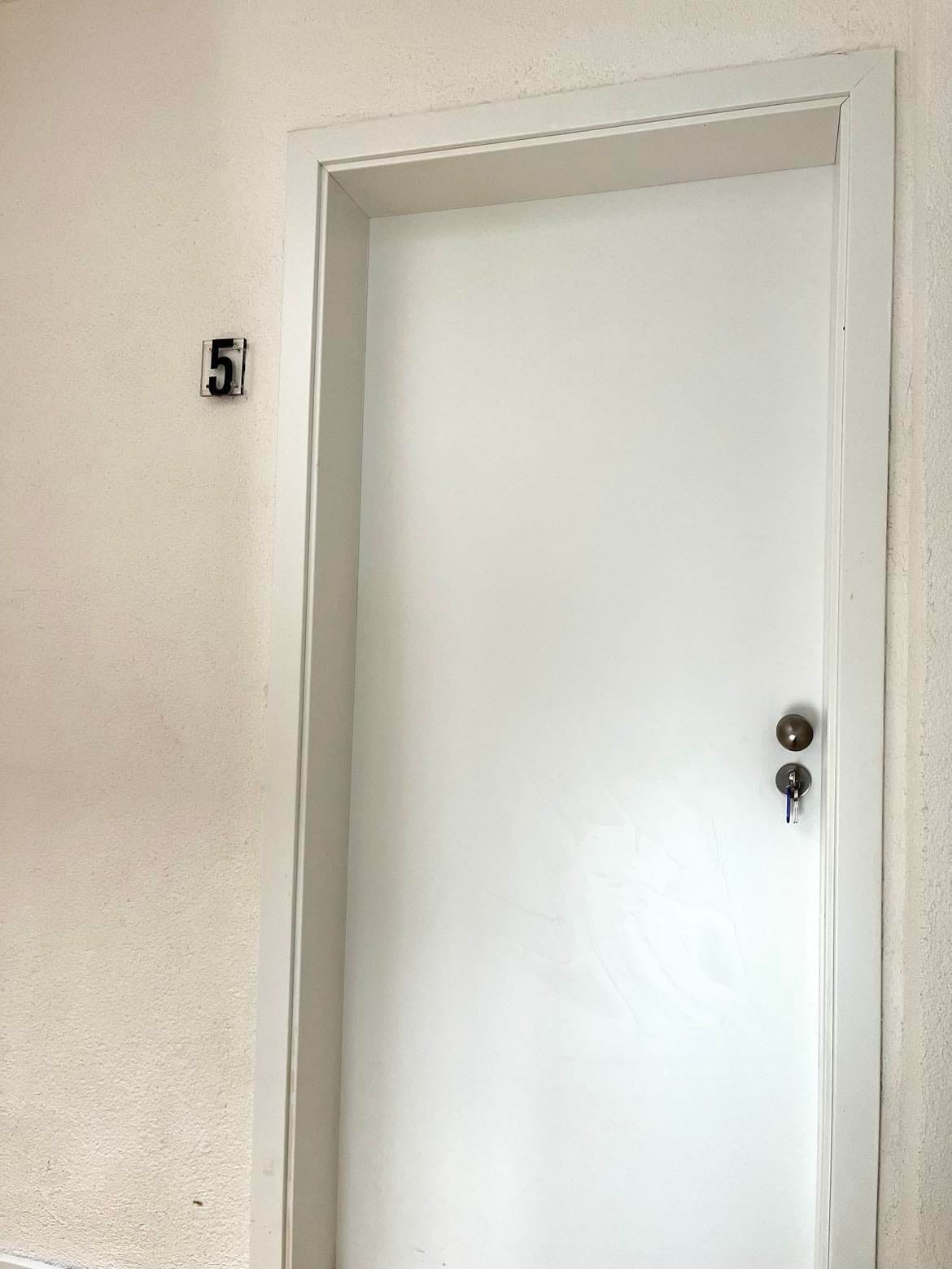 A white door is positioned in the center of the frame, with a visible door handle and keyhole. The number '5' is displayed prominently on the wall next to the door, indicating the apartment number. The surrounding wall is a neutral tone.