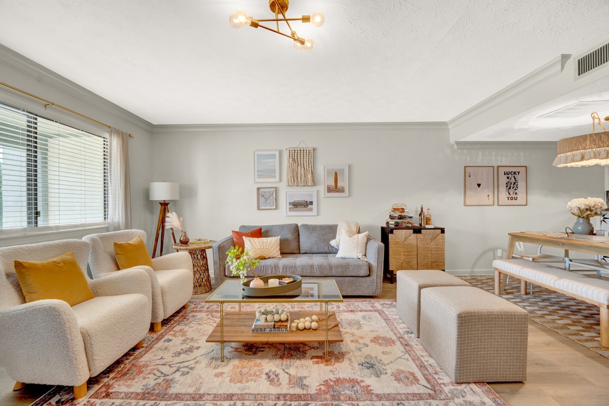 A bright living room features a mix of comfortable seating, including two armchairs and a gray sofa. A glass coffee table is placed atop an area rug, showcasing decorative items. Natural light filters through large windows, enhancing the warm color palette and inviting atmosphere.
