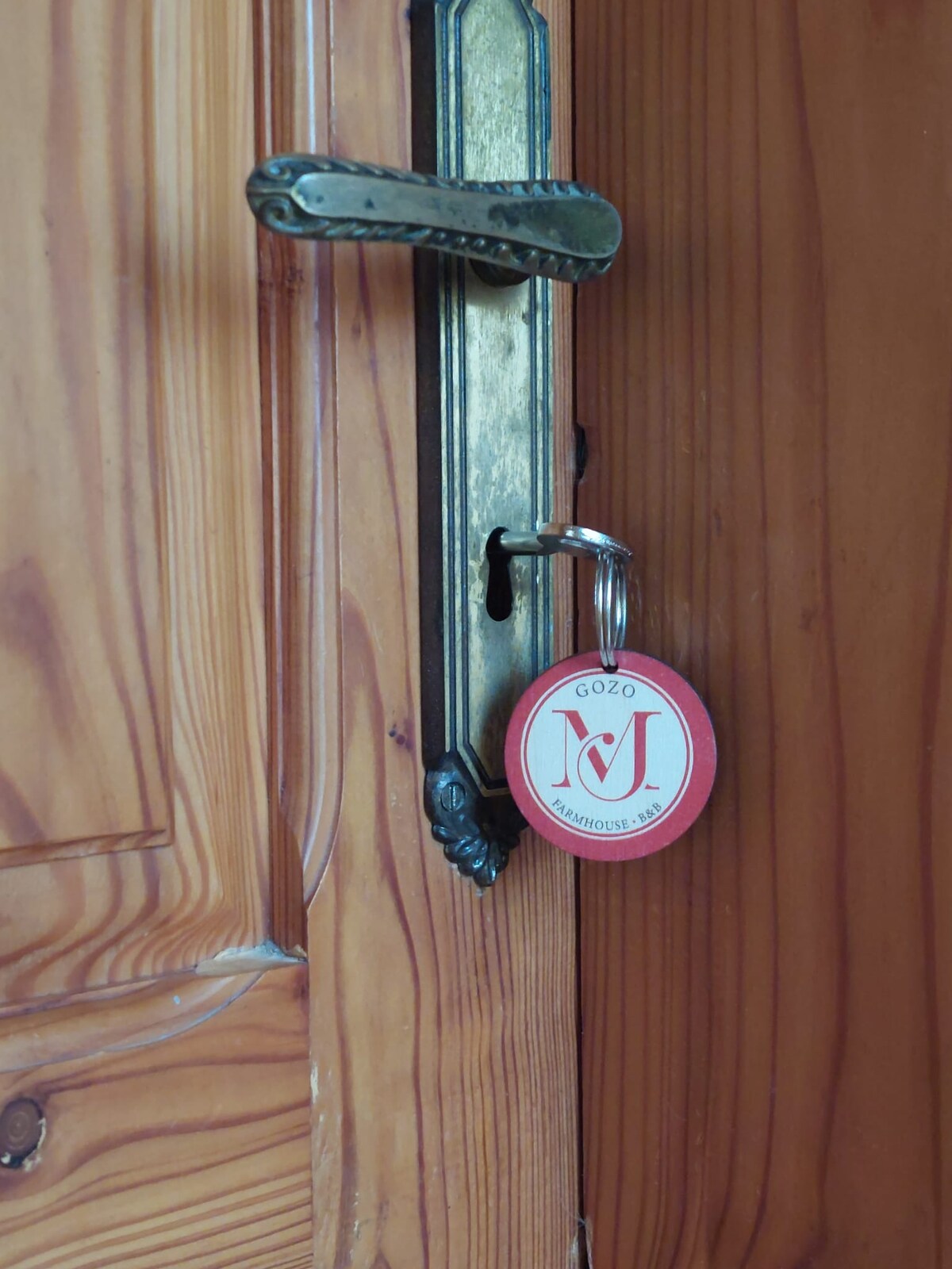 A close-up view of a wooden door featuring a classic latch mechanism. A key with a round red tag displaying the name 'MJ' is prominently visible, adding a personal touch to the entrance.