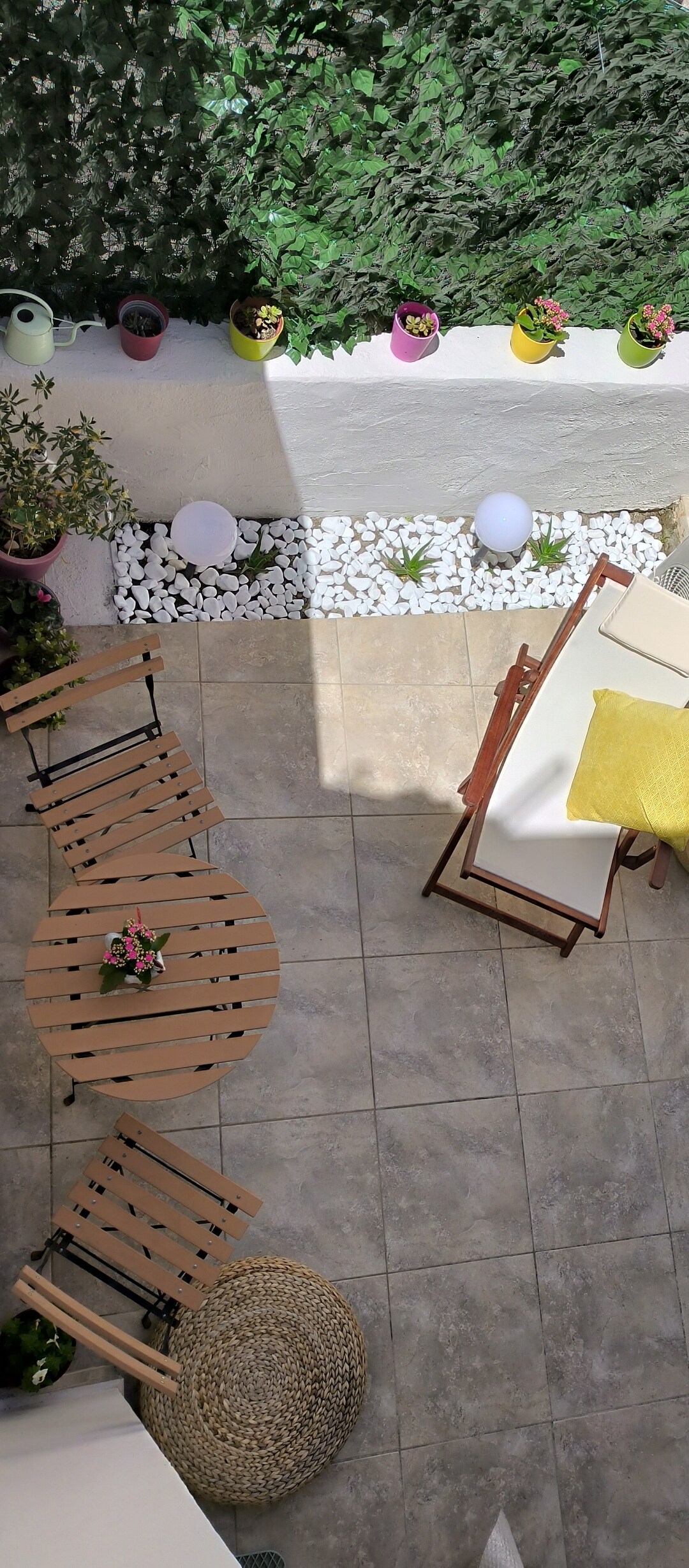 An outdoor courtyard features a small wooden table with two matching folding chairs beside it. The area is adorned with potted plants and white pebbles, complemented by a soft light source. A larger table with a yellow accent pillow invites relaxation.
