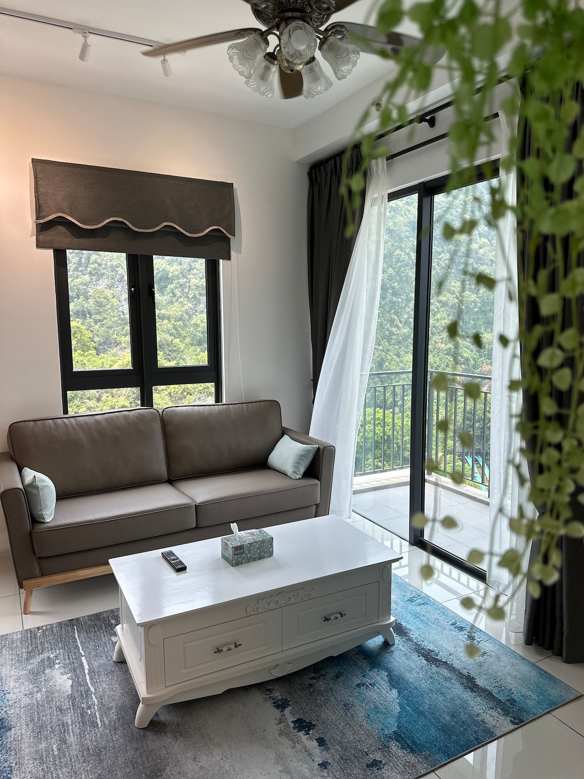 A modern living area showcases a beige sofa accented with two light blue cushions, positioned in front of a set of tall glass doors. A white coffee table is centered on a blue patterned rug. Natural light fills the space from the balcony overlooking lush greenery.