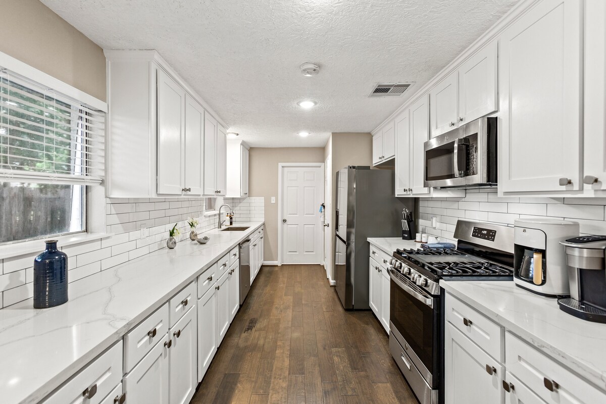 A modern kitchen is presented with white cabinetry and sleek quartz countertops. Appliances include a stainless-steel refrigerator and oven. Light-colored backsplash tiles complement the spacious layout, which features ample counter space and a window providing natural light.