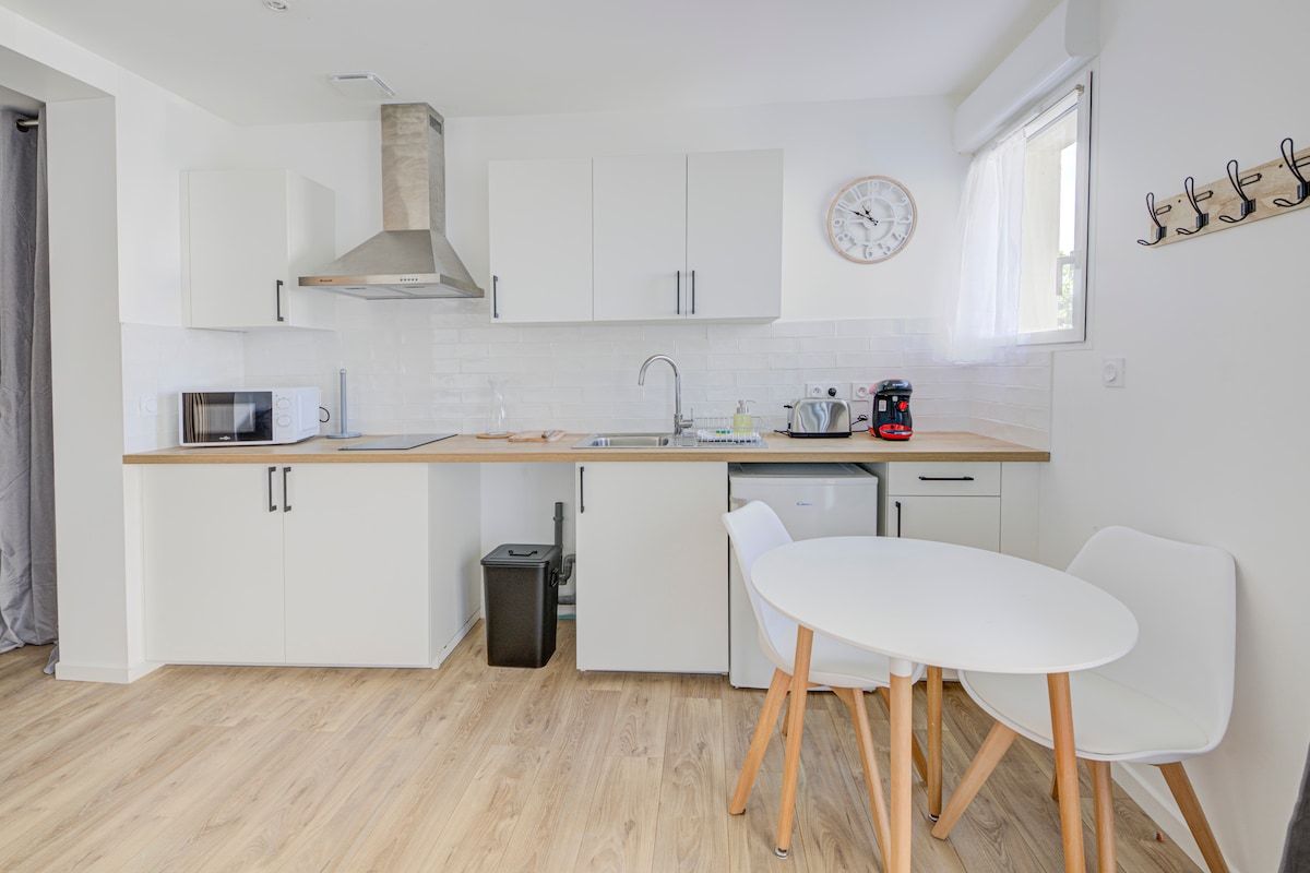 A functional kitchen area features modern white cabinetry, complemented by a wooden countertop. Stainless steel appliances, including a microwave and coffee maker, are visible. A round dining table with two white chairs stands nearby, with a small black trash bin discreetly placed under the counter.