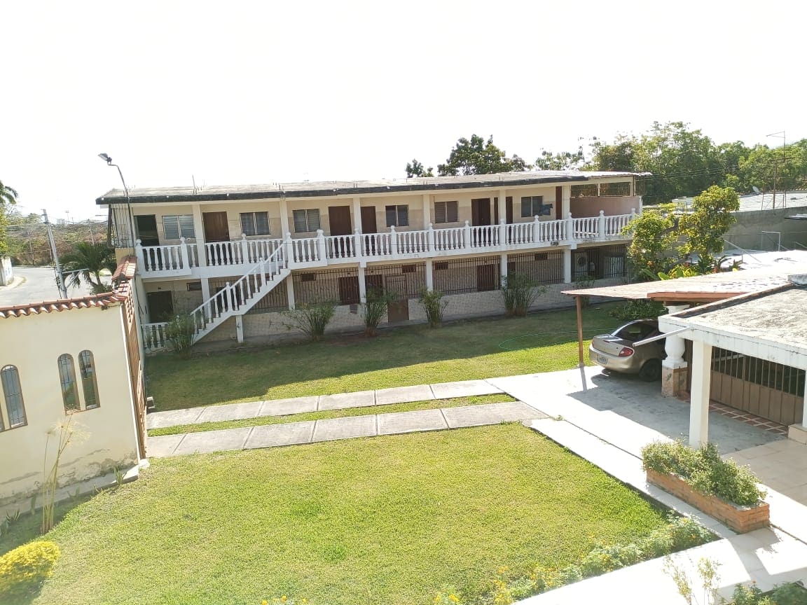 A two-story building is visible, featuring a series of individual rooms with balconies. A grassy area in the foreground adds greenery to the scene. Parking space is located adjacent to the structure, with a roadway leading to the entrance.