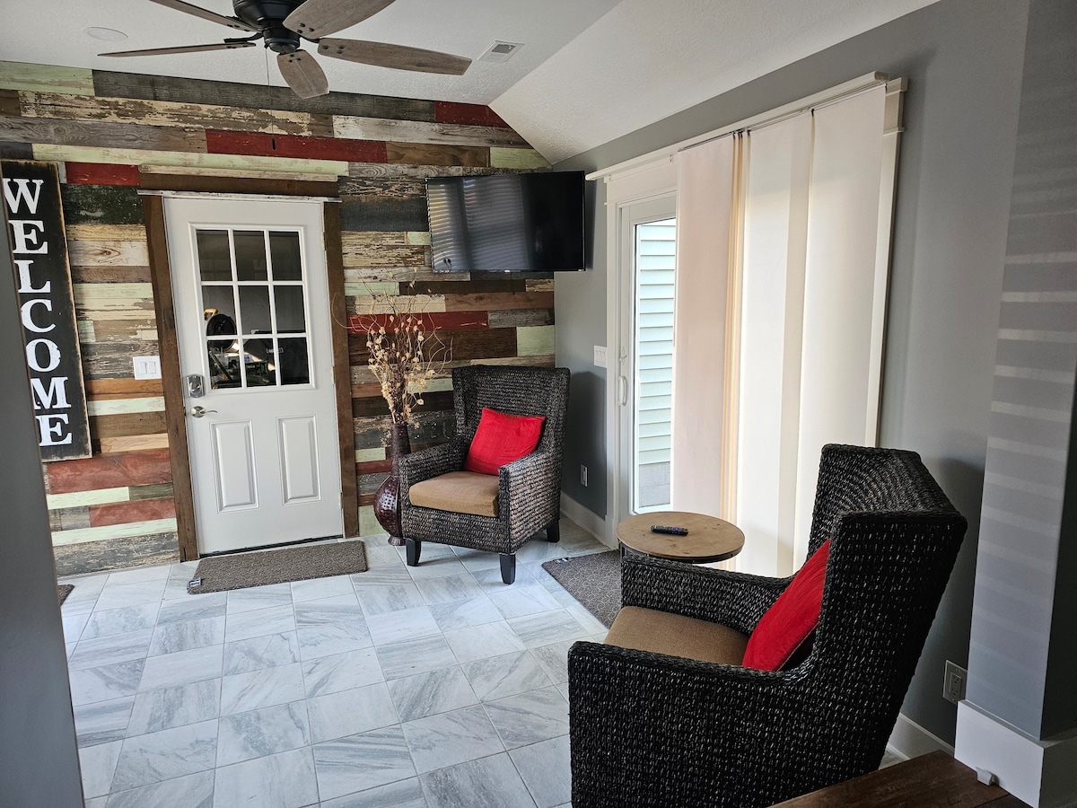 A welcoming entryway features a wooden door with a glass panel, flanked by a textured accent wall made of reclaimed wood. Two comfortable chairs with red cushions are positioned beside a small table, and a TV is mounted above for added convenience.