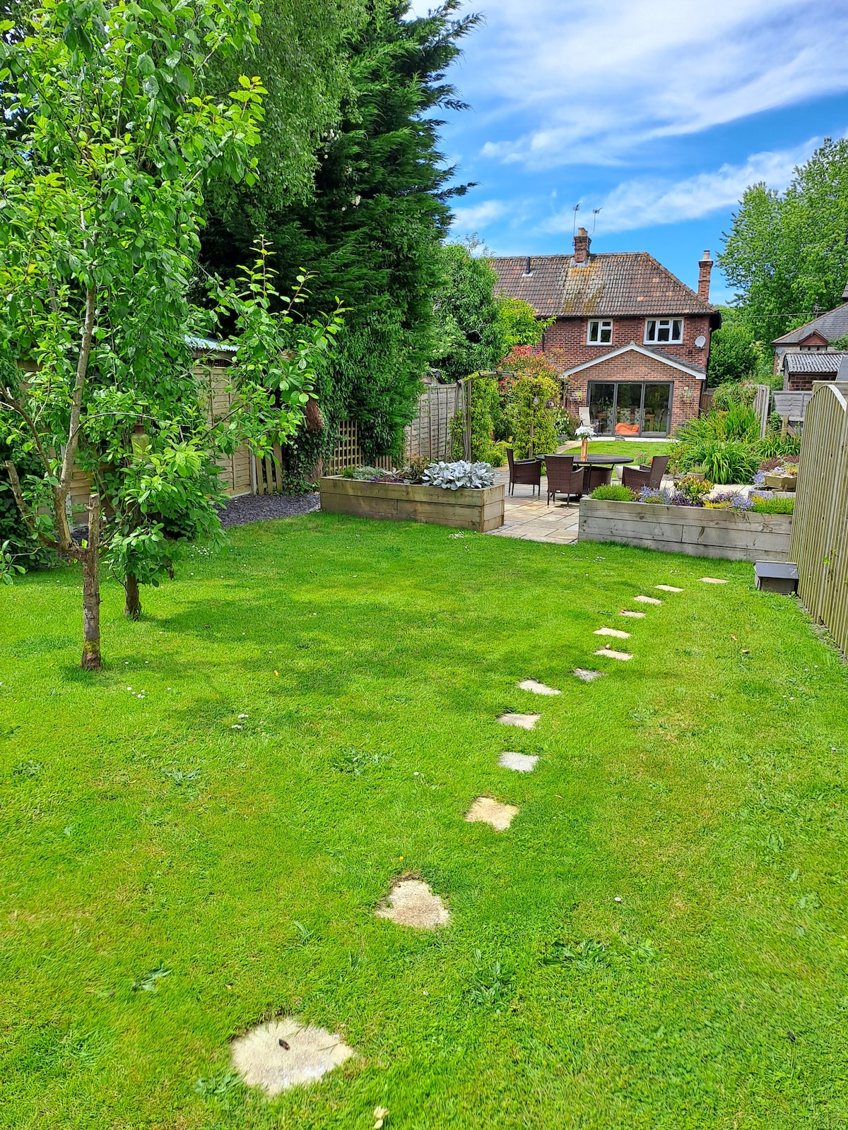 A spacious garden area features a well-maintained lawn with stepping stones leading to a patio seating area. Lush green trees are visible, along with flower beds, providing a serene outdoor setting. The house stands in the background, framed by the greenery.