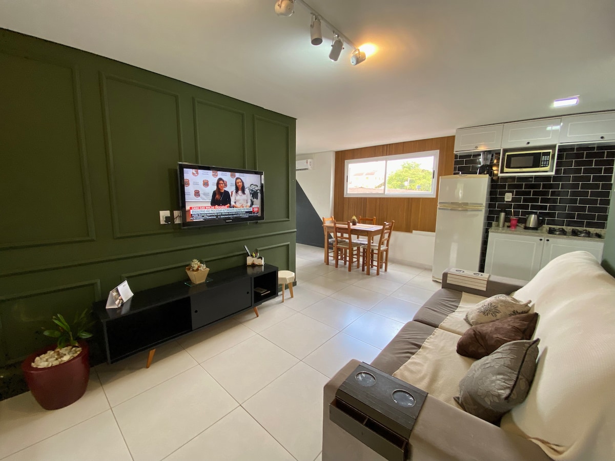 A cozy living area features a comfortable sofa accompanied by decorative cushions. A flat-screen television is mounted on a dark media unit. Natural light floods in from a window, illuminating a dining table for four. The kitchen area is visible in the background.