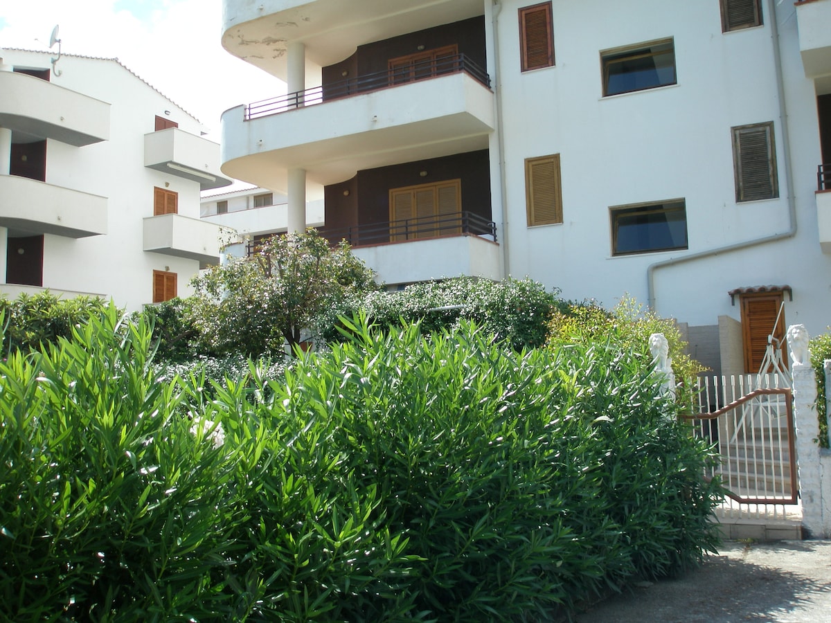 A multi-level building is surrounded by lush greenery and flowering plants. The entrance is marked by a white gate leading to a pathway. Balconies with wooden railings are visible on the upper floors, and windows are adorned with wooden shutters.