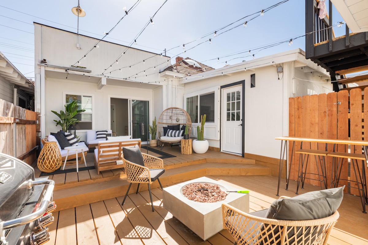 An outdoor patio area features a spacious wooden deck adorned with string lights. Various seating options include a cozy hanging chair, a sofa, and stylish lounge chairs. A fire pit sits at the center, complemented by green plants and a grill on one side.