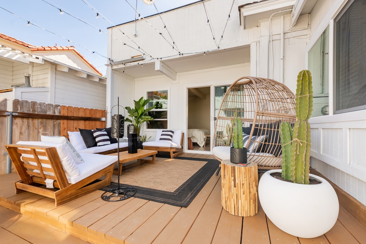 A private outdoor patio features a lounging area with wooden chairs and a cozy nest swing. Potted plants add a touch of greenery, while comfortable cushions enhance the seating arrangement. A softly textured rug defines the space, providing a welcoming atmosphere for relaxation.