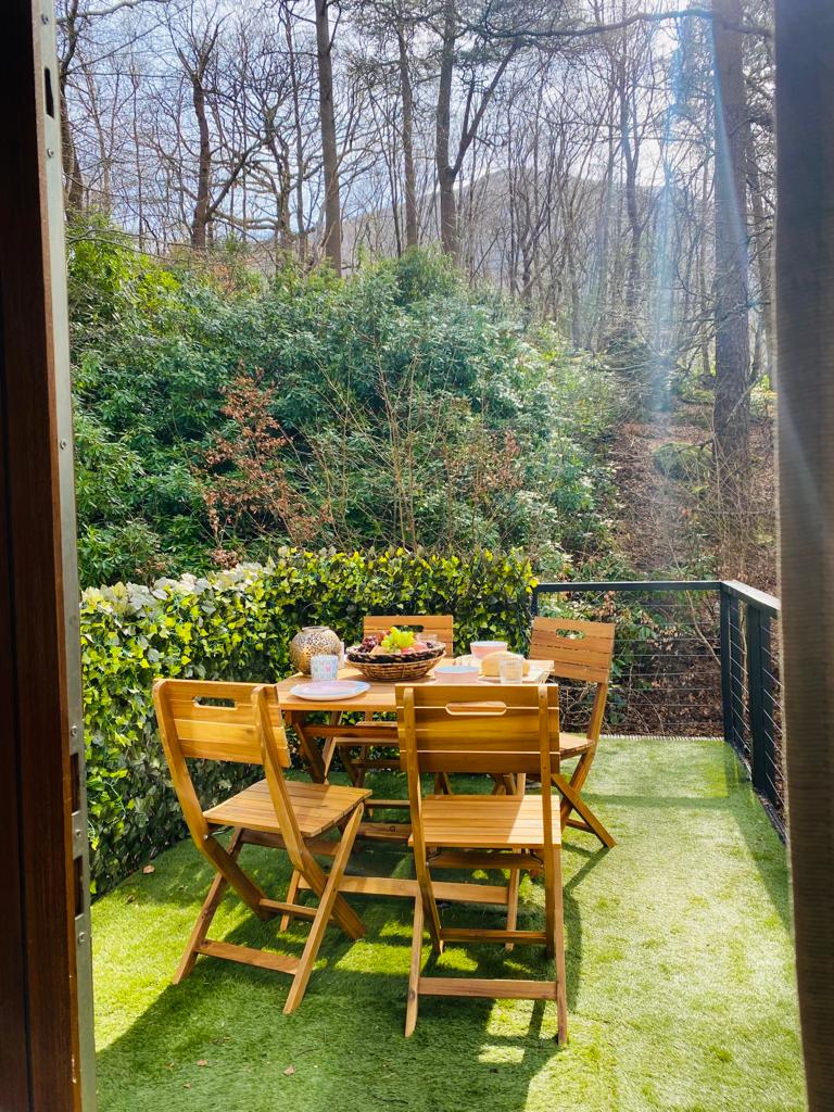 An outdoor seating area is presented, featuring a wooden table surrounded by four folding chairs. The space is adorned with lush greenery in the background, while faux grass covers the floor. Morning sunlight filters down, creating a serene ambiance.