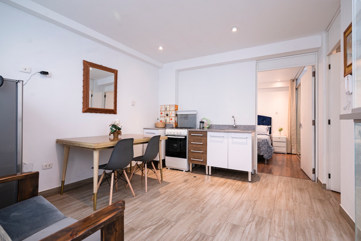 A modern kitchen area includes an integrated stove and refrigerator, with a table set for four. Light-colored cabinetry complements the open layout, which is well-lit by natural light. A mirror on the wall enhances the spacious feel, leading to a cozy sleeping area in the background.