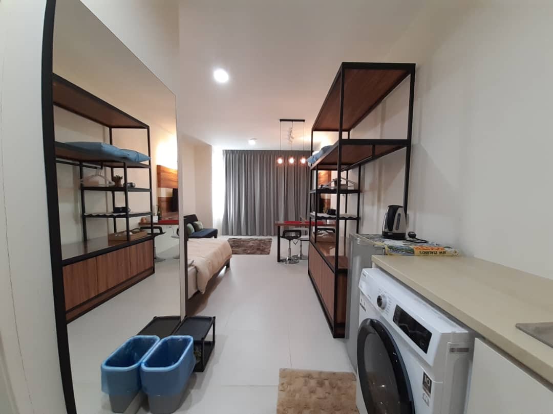 A modern interior is observed, featuring a spacious layout accented by contemporary shelving units. A washing machine is positioned near a countertop. A large mirror reflects the living space, which includes a bed and soft furnishings in muted tones. Natural light filters through the floor-to-ceiling curtains.