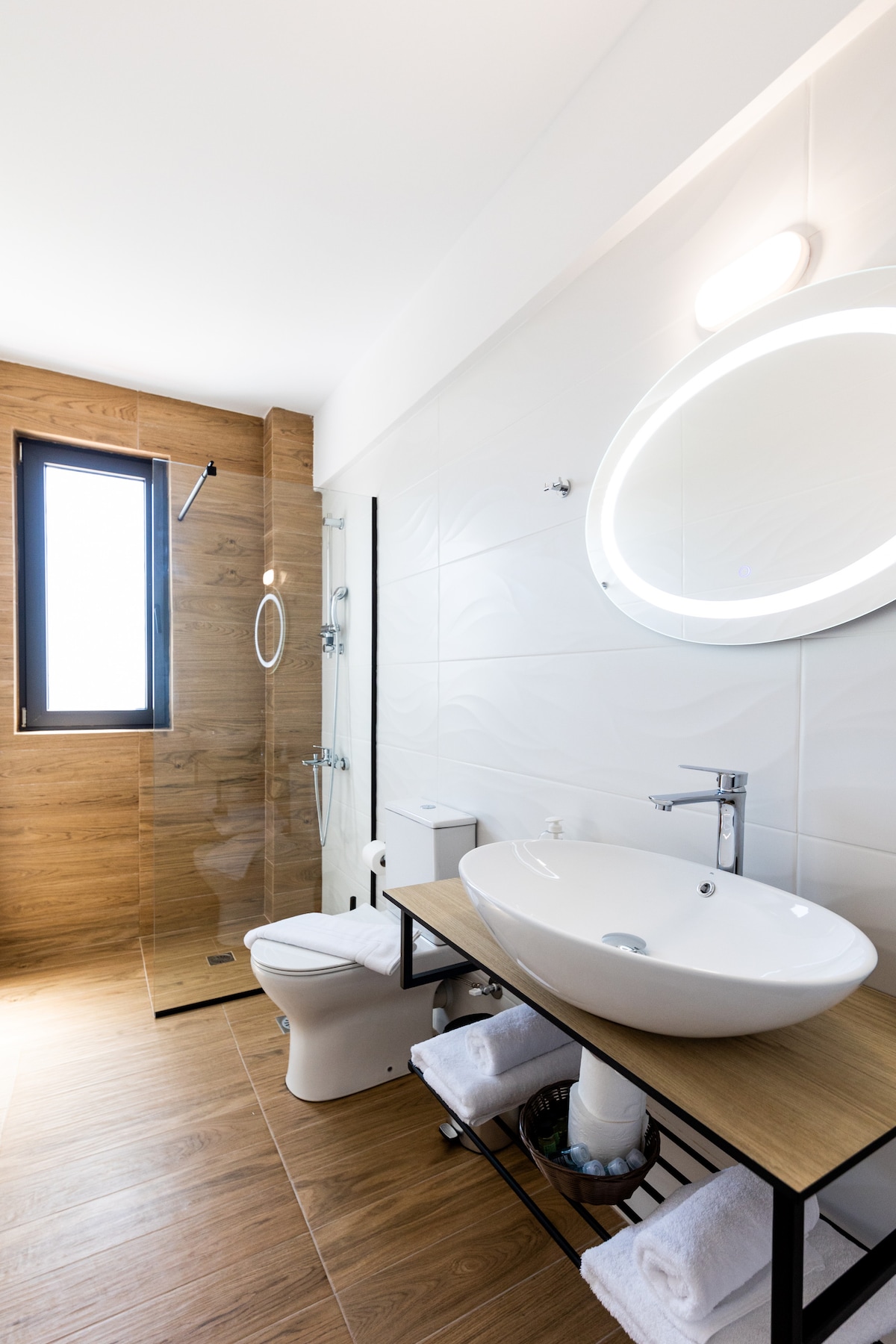 A modern bathroom features a wall-mounted sink with a round mirror illuminated by soft lighting. A walk-in shower with glass panels is visible, while natural light enters through a narrow window. Grab bars and neutral-colored towels provide practical elements in the space.