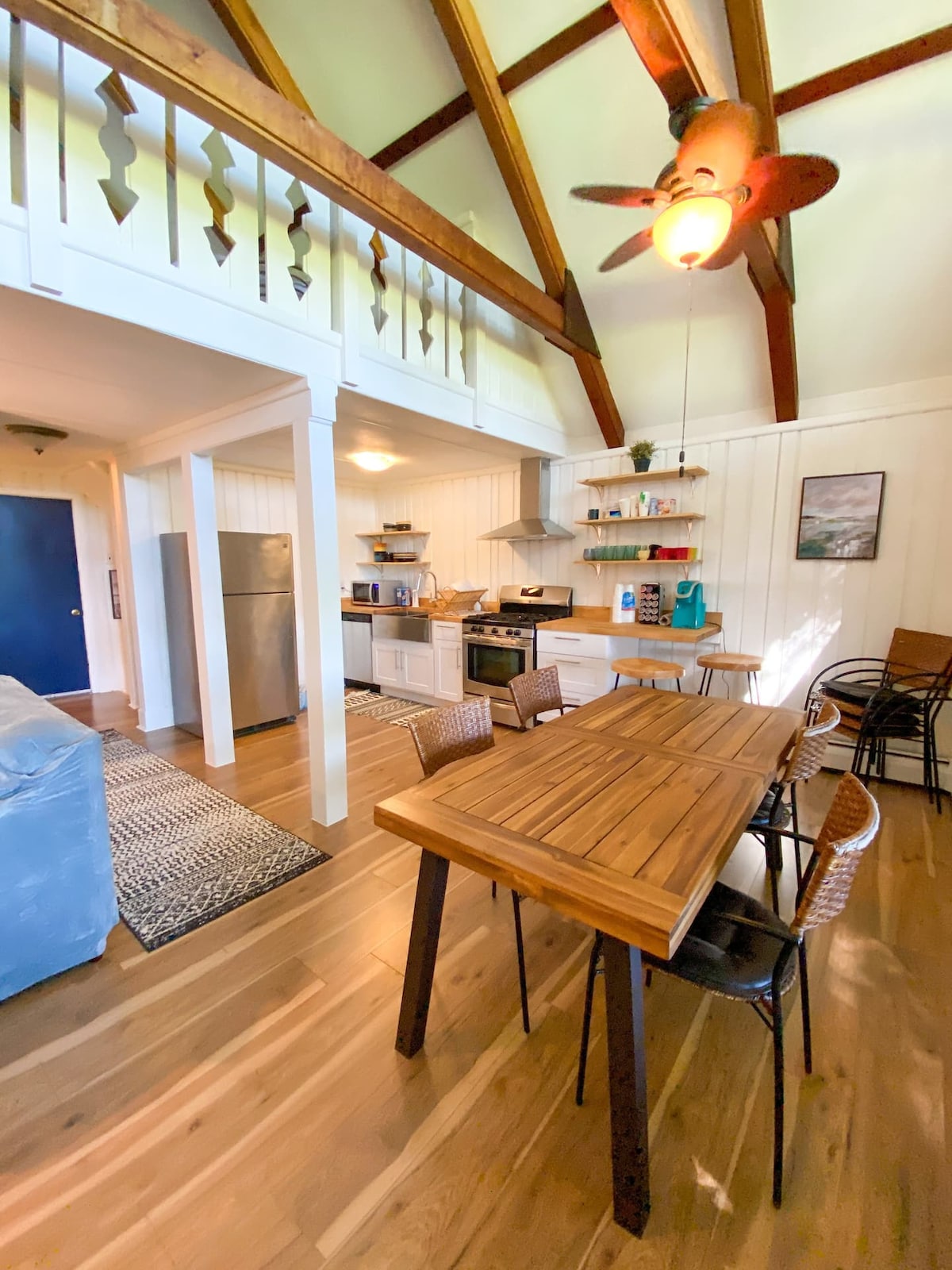 The kitchen and dining area feature a wooden table with seating for six, positioned beneath a sloped ceiling with exposed beams. Modern appliances, including a refrigerator and oven, are visible in the fully equipped kitchen. Natural light brightens the space through a nearby window.