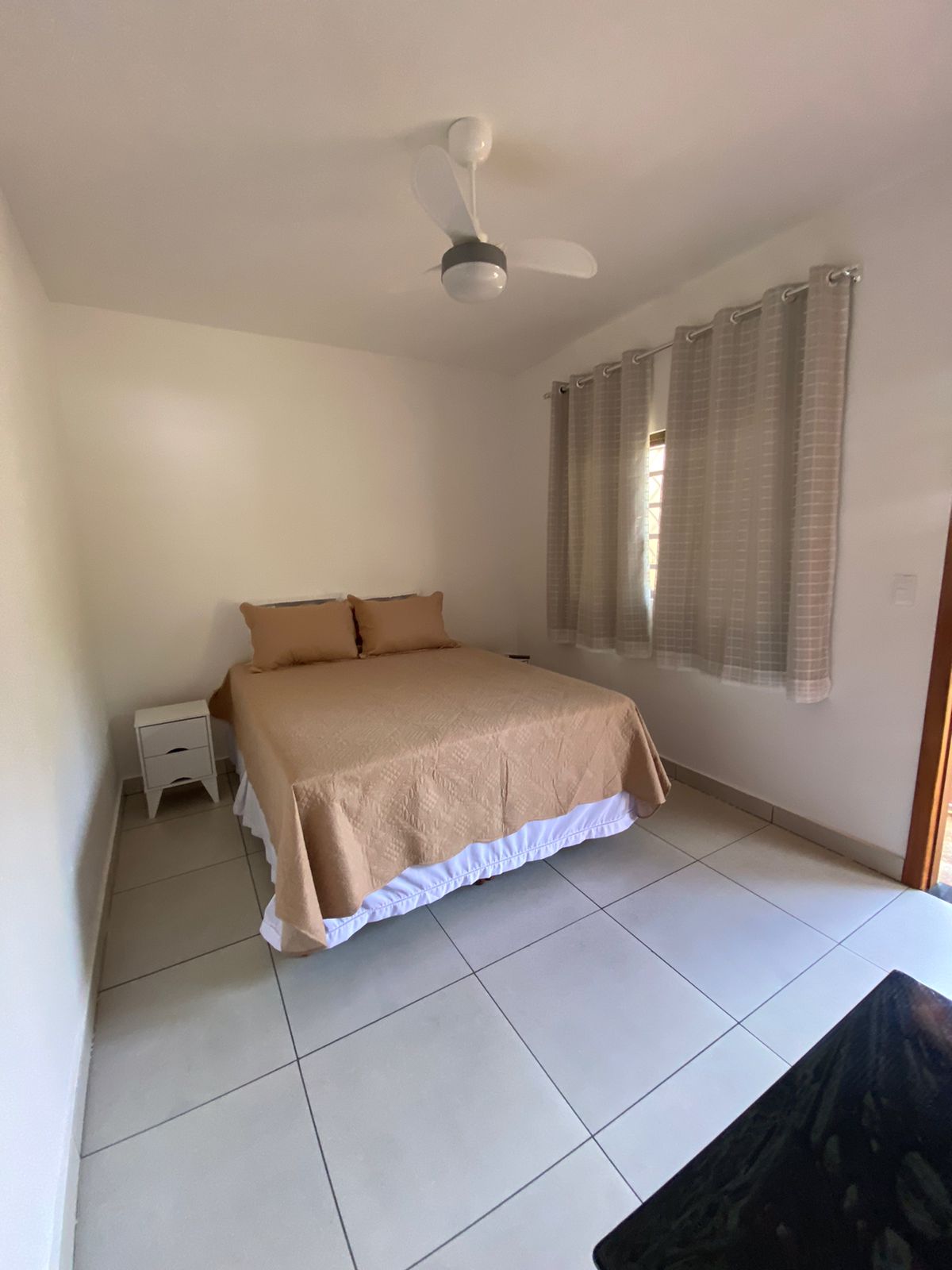 A cozy sleeping area features a spacious bed with neutral-toned bedding and two matching pillows. Natural light enters through a partially open window, complemented by light, sheer curtains. A small bedside table is positioned next to the bed, enhancing the simplicity of the room.