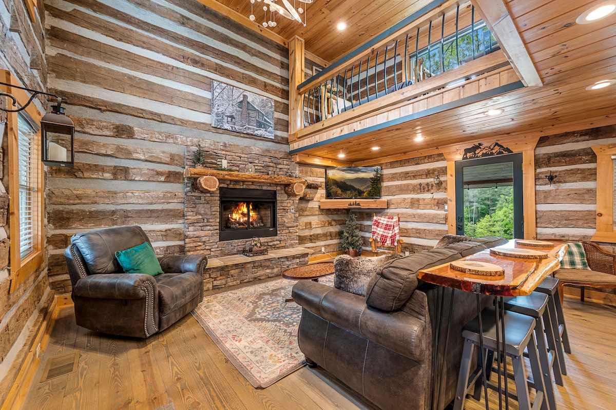A spacious living area features rustic log walls and a stacked stone fireplace, creating a cozy centerpiece. Comfortable seating is arranged around the fireplace, complemented by a wooden dining table nearby. Windows allow natural light to illuminate the space, enhancing the warm wood tones.