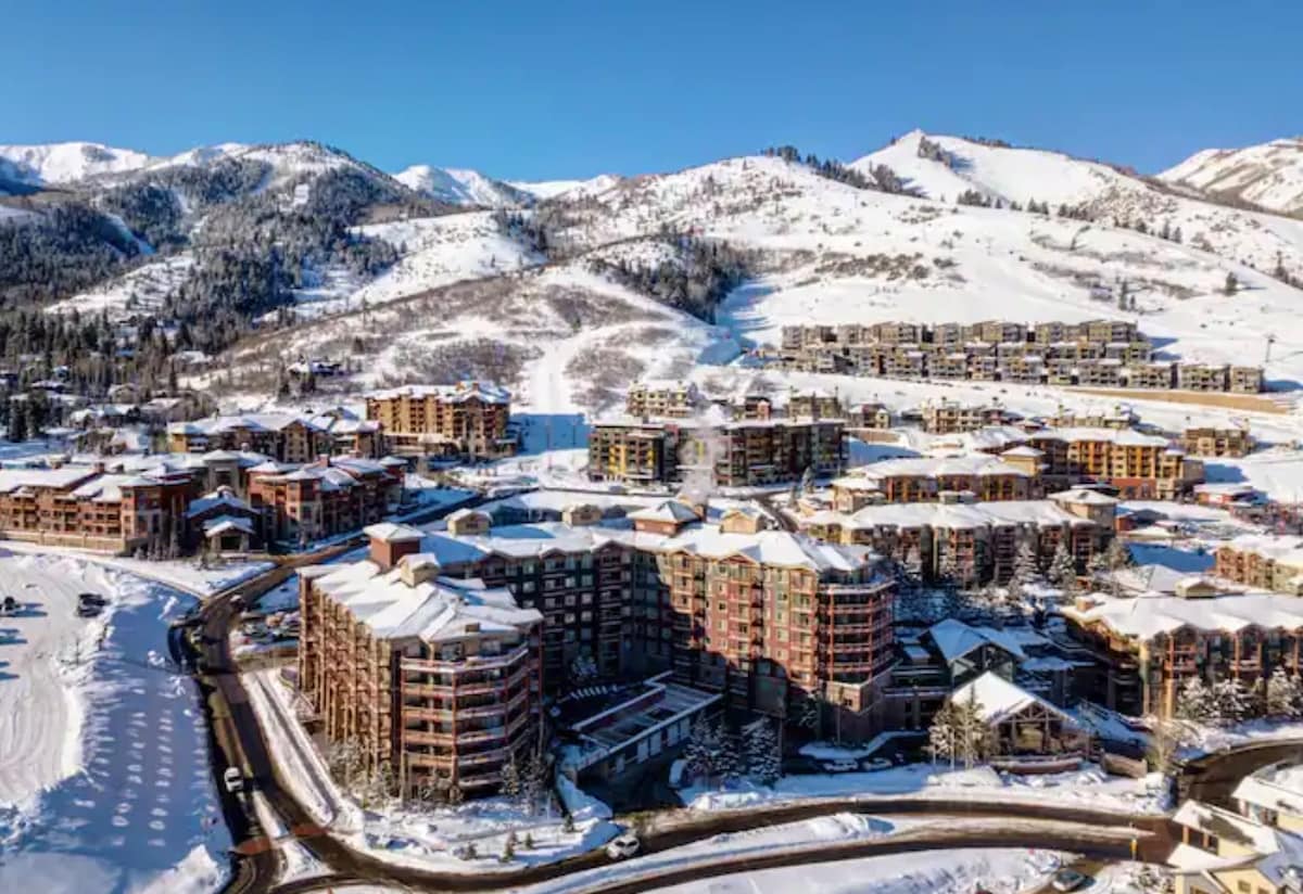An aerial view captures the Westgate Park City Resort & Spa nestled amidst snow-covered mountains. Buildings with a blend of warm colors and snowy roofs are presented, showcasing the expansive layout of the resort. The surrounding landscape highlights serene slopes, indicative of the winter season.
