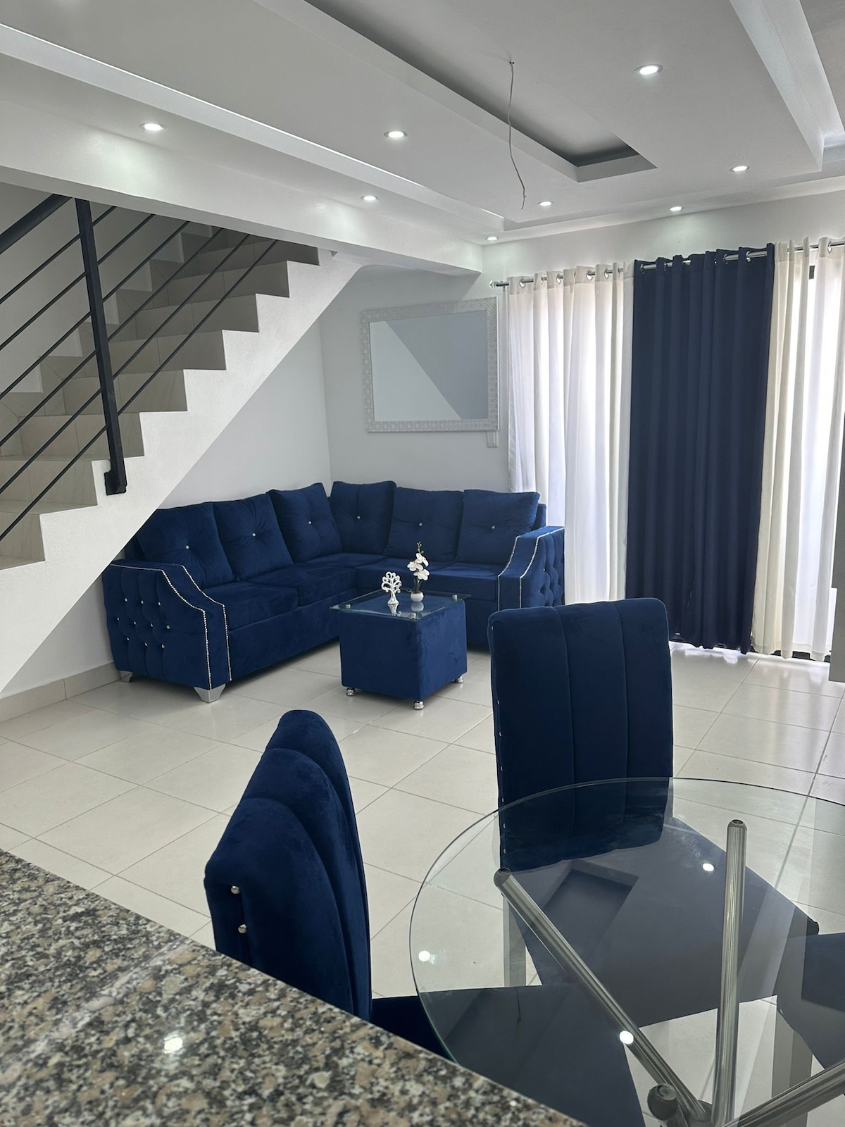 A modern living area features a deep blue sectional sofa arranged with coordinating accent chairs. A glass table is positioned nearby, surrounded by upholstered chairs. Natural light filters through sheer and curtain panels, illuminating the neutral tile flooring.