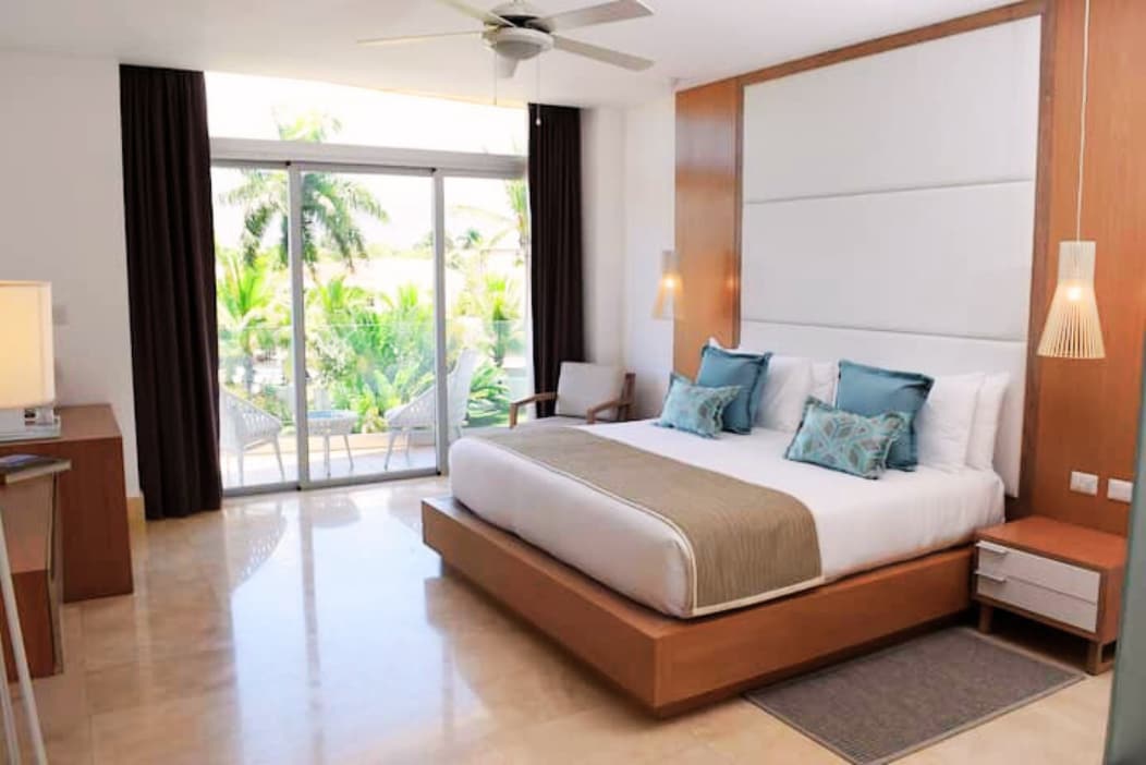 A spacious bedroom features a large bed with decorative pillows, complemented by a soft-toned bedspread. Light floods in through sliding glass doors that open to a patio surrounded by tropical foliage, while a ceiling fan and modern fixtures enhance the room's comfort.