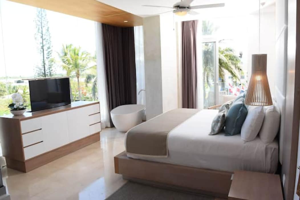 A well-appointed bedroom suite features a spacious bed with light bedding and decorative pillows. Natural light fills the room through expansive windows, offering views of palm trees outside. A modern television is positioned on a sleek dresser, while a freestanding tub adds an element of relaxation.