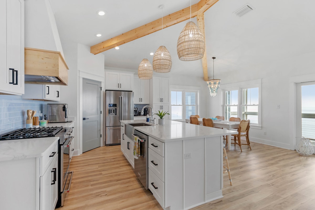 A modern kitchen is highlighted with sleek white cabinetry and stainless steel appliances. A large island with a marble countertop provides ample space for meal preparation. Three woven pendant lights hang above, adding a touch of warmth, while large windows offer views of the Gulf.