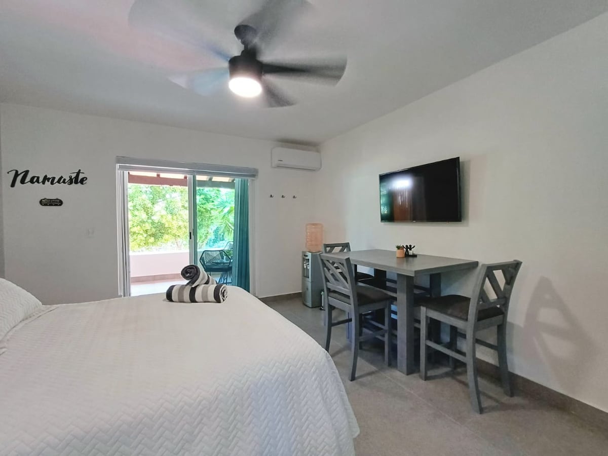 The loft-style accommodation features a spacious bed with smooth linens, complemented by a modern dining table with four chairs. A wall-mounted television is visible, along with light streaming in from a sliding glass door that leads to a balcony area.