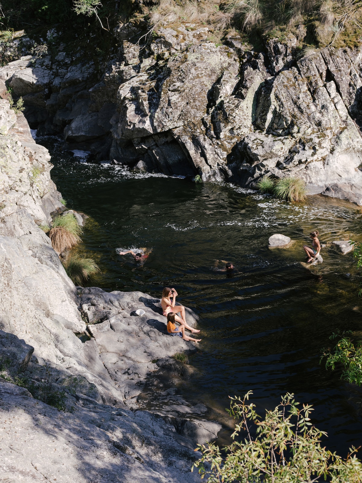 A natural swimming area is seen, surrounded by rocky formations and lush greenery. Several individuals relax in the clear water, while some sit on the smooth stones along the shore. A calm stream flows gently through the landscape, creating a serene outdoor setting.