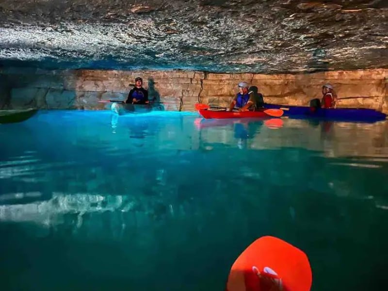 A serene underground cave is depicted, with calm waters reflecting natural stone walls. People are seen in colorful kayaks, paddling or resting. Soft light enhances the tranquil atmosphere as natural formations create a captivating backdrop.