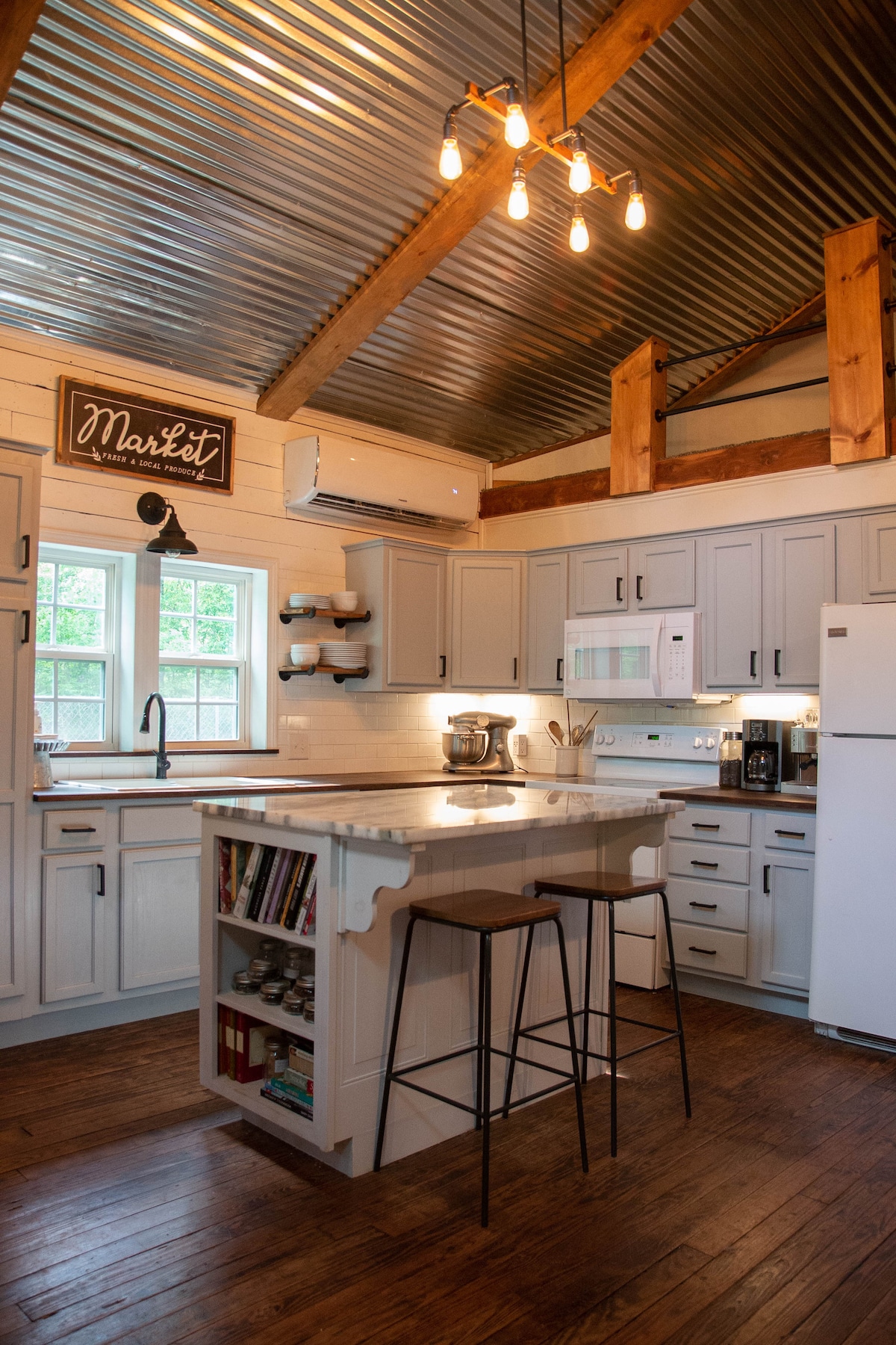 The kitchen features modern appliances with a central island that includes seating. Light wooden cabinetry is complemented by a corrugated metal ceiling and warm overhead lighting. Windows provide natural light, enhancing the functional layout for cooking and gathering.
