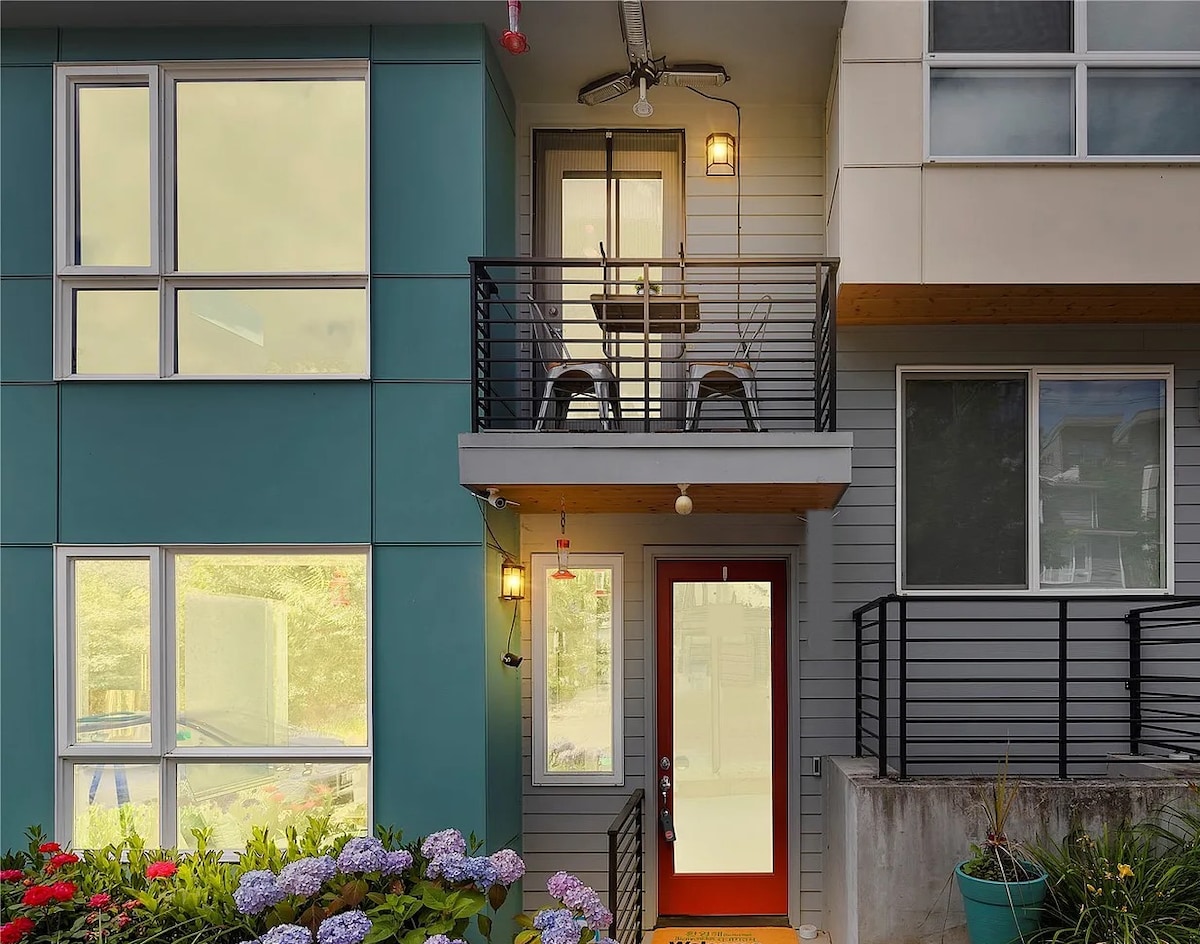 A modern exterior features a private entrance with a bold red door, flanked by large windows. A small balcony with two chairs is visible, surrounded by vibrant flowers and greenery, emphasizing a welcoming outdoor space.