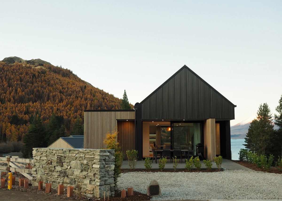 The architectural exterior of the home features a modern design with a combination of wood and metal finishes. Large windows reveal glimpses of the interior, while landscaped greenery enhances the entryway. The backdrop showcases rolling hills and Lake Tekapo, creating a serene atmosphere.