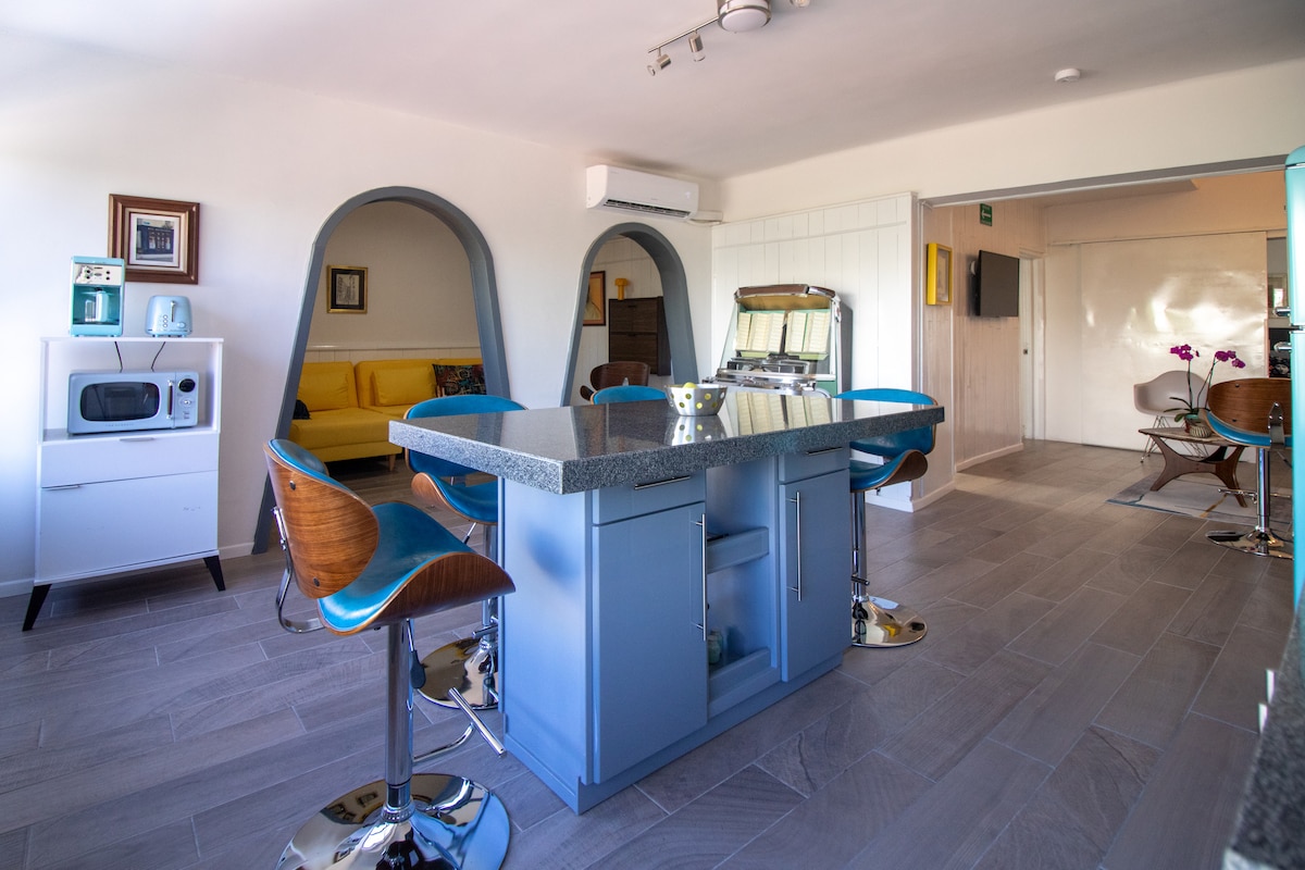 A stylish kitchen area features a modern island with bar stools, surrounded by arched openings that lead to a cozy living area. Essential appliances are available, including a microwave. Large windows allow natural light to brighten the space, contributing to an inviting atmosphere.