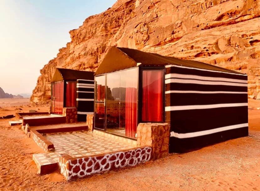 Two cabins with glass-fronted facades are set against a backdrop of towering, rocky cliffs. The structures feature black and white striped fabric, complemented by red curtains. A stone pathway leads to the cabins, which are surrounded by the sandy desert landscape.