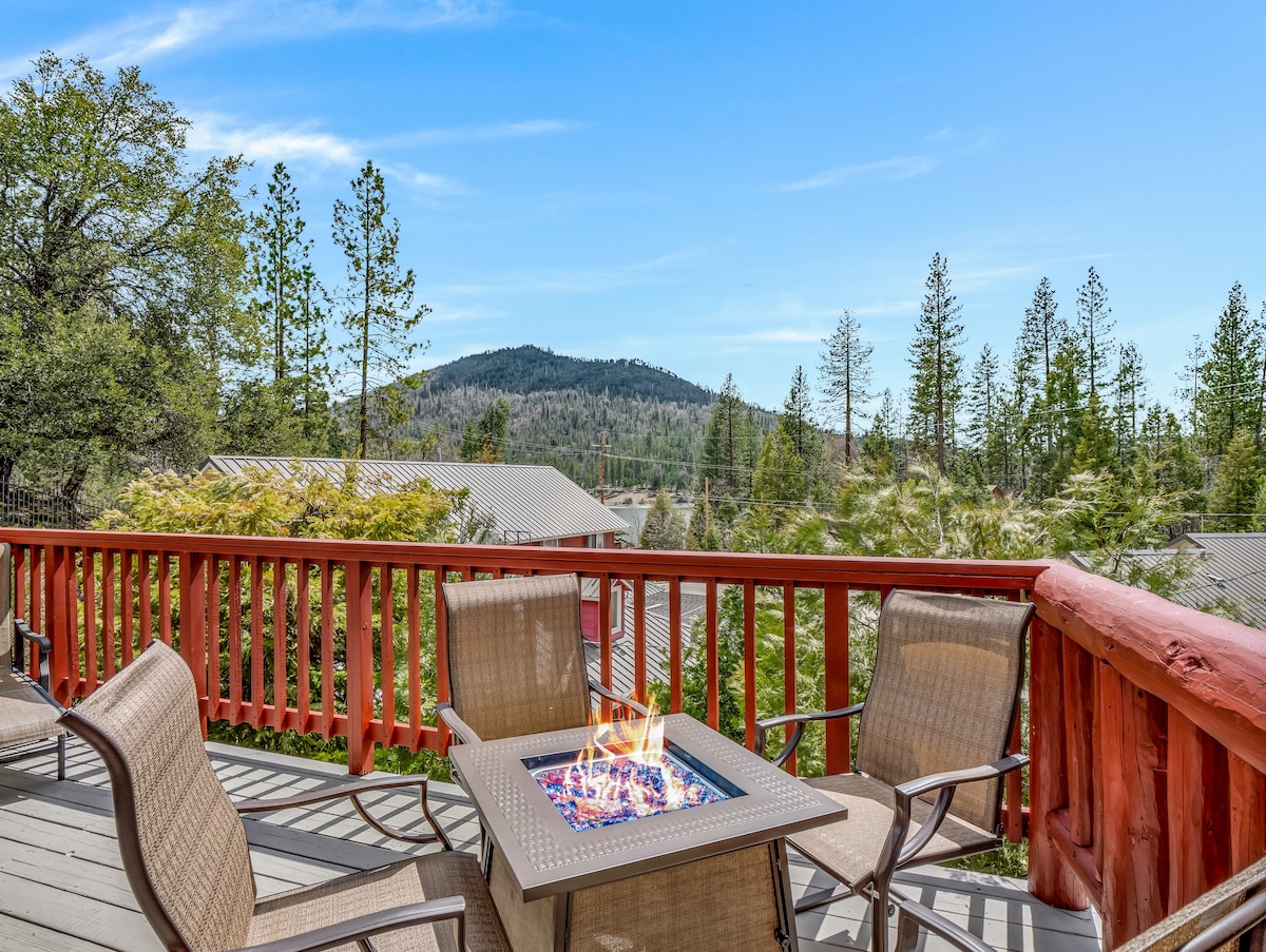 An inviting outdoor space features a patio table with a fire pit, surrounded by cozy chairs. The view encompasses lush trees and distant mountain ranges under a clear blue sky, offering a peaceful setting for relaxation.