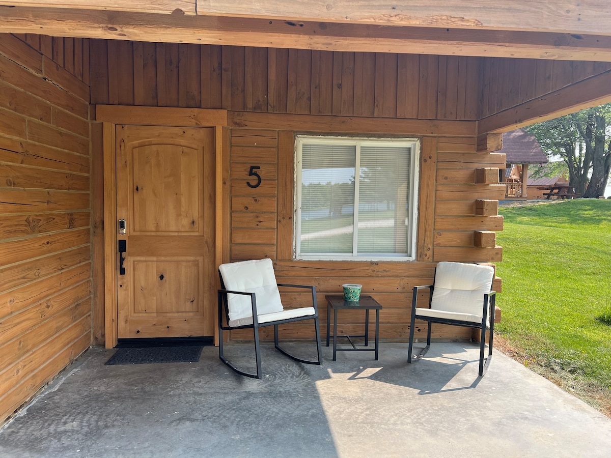 The porch area of a rustic log cabin features two white chairs positioned in front of a wooden door. A small potted plant sits on a side table between the chairs. A vast grassy area and trees are visible in the background.