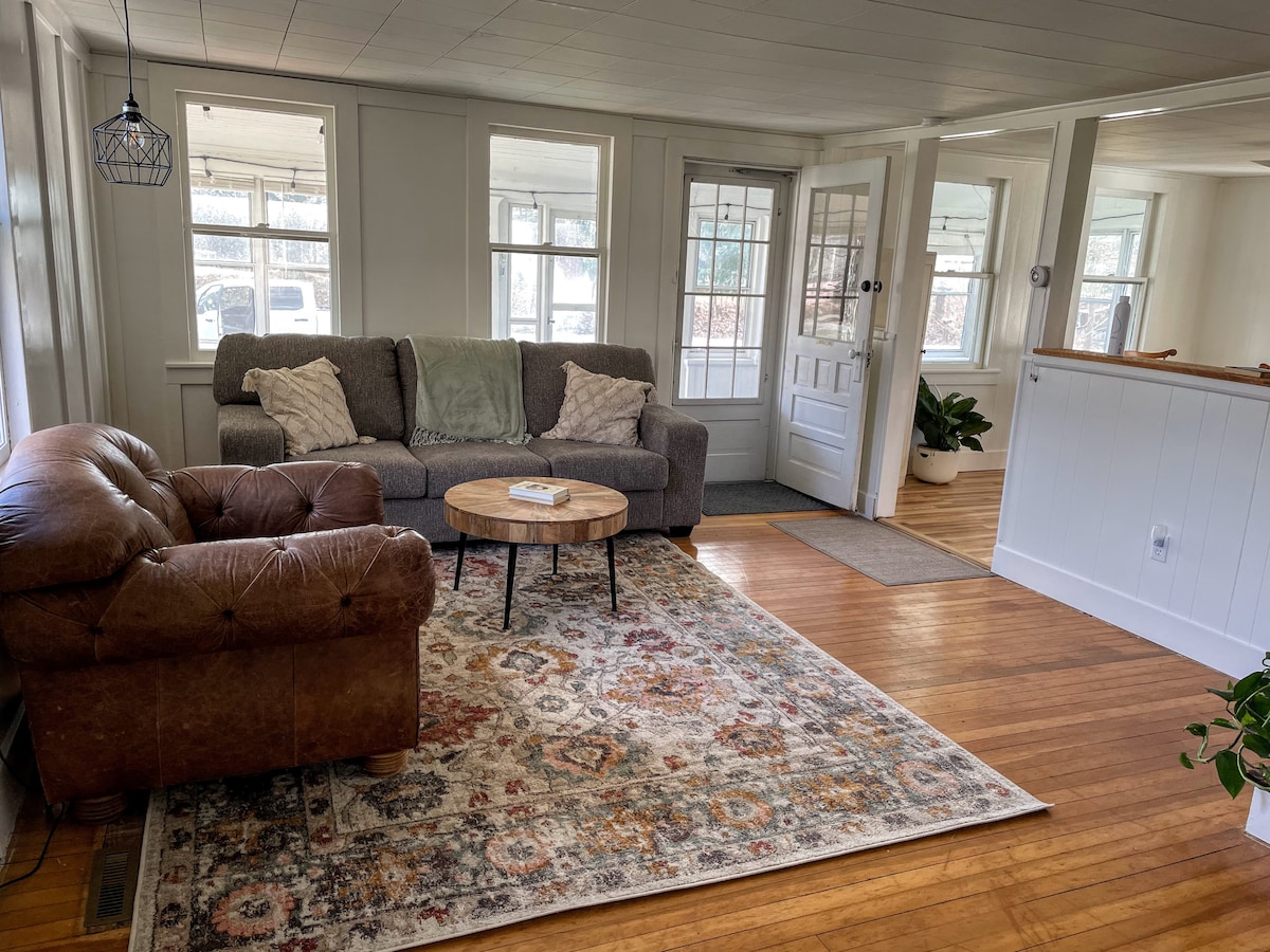 A bright living space features a large gray sofa and a cozy brown armchair paired with a round wooden coffee table. A colorful area rug lies on polished wooden floors, while natural light filters through multiple windows, enhancing the welcoming atmosphere.