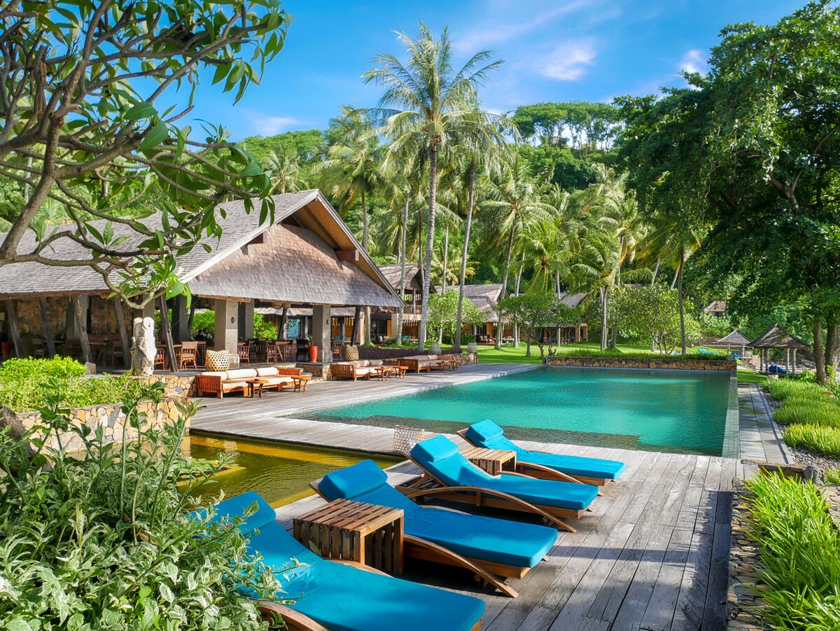 An inviting outdoor area features a tranquil infinity pool surrounded by lush greenery and swaying palm trees. Comfortable sun loungers with vibrant blue cushions line the pool deck, providing a perfect spot for relaxation and sunbathing amid the serene landscape.