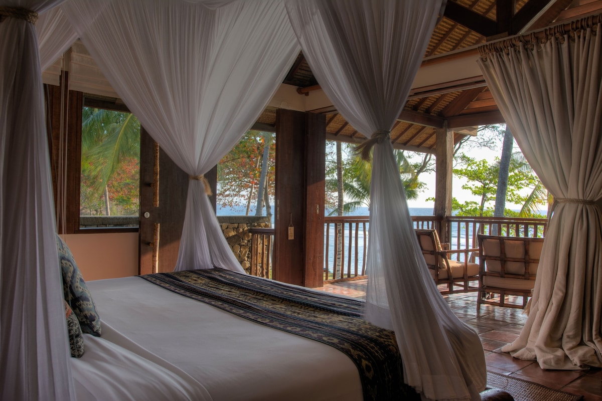 A spacious suite features a king-sized bed draped with sheer white curtains, offering views through large open doors. Natural light floods the room, illuminating wooden accents and a textile throw. A private terrace with seating overlooks the lush surroundings and ocean beyond.
