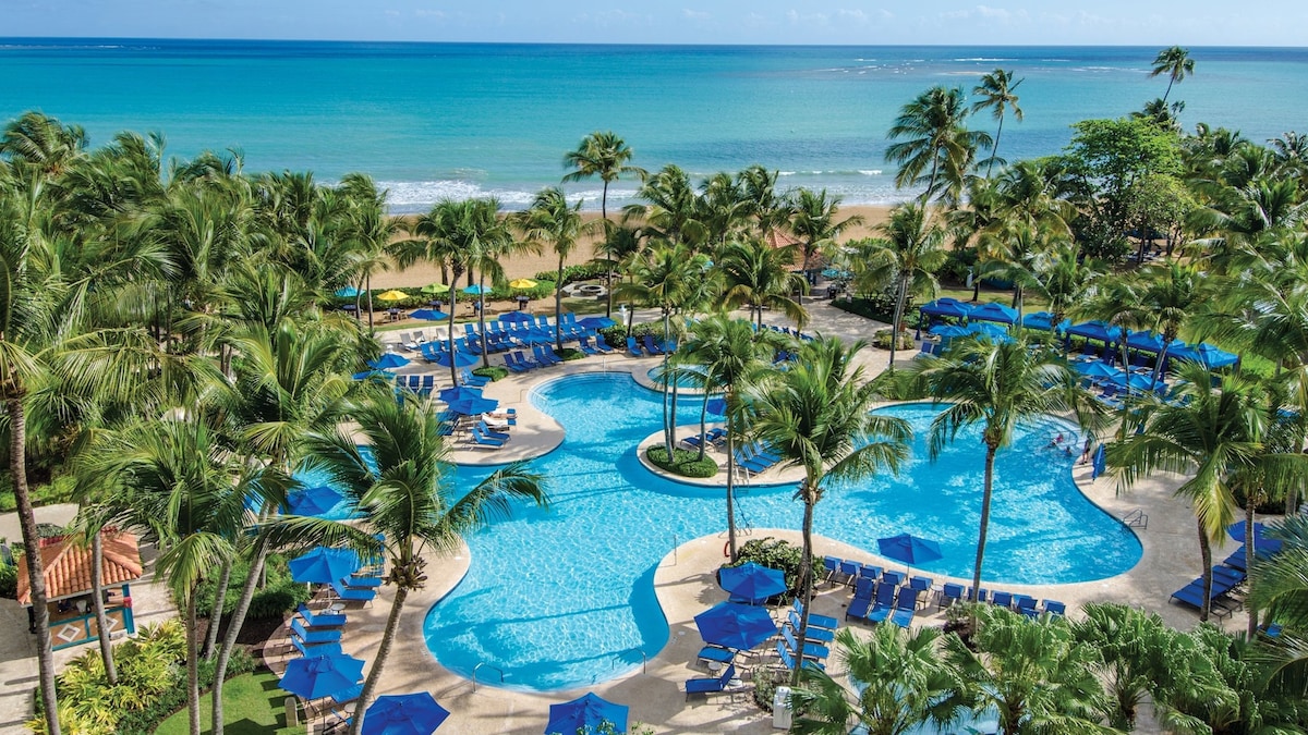 A spacious pool area features multiple connected swimming pools surrounded by palm trees and beach chairs. Bright blue umbrellas provide shade for guests lounging beside the water, while the Atlantic Ocean and golden sand beach are visible in the background under a clear sky.