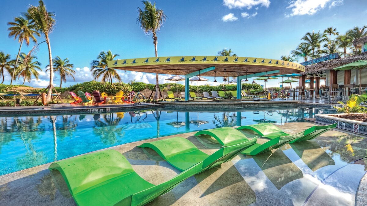 A large swimming pool features vibrant green lounge chairs positioned along the water's edge. An open-air cabana with a colorful roof provides shade, with palm trees swaying in the background. Clear blue skies and tropical greenery surround the area, creating a relaxing atmosphere.