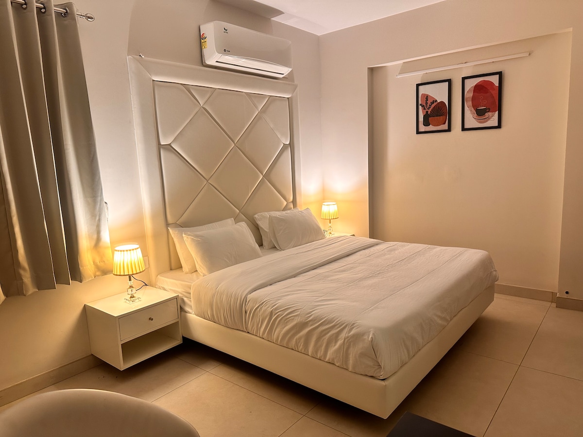 A spacious bedroom features a large upholstered headboard and a comfortable bed. Two bedside tables with lamps provide soft lighting. Light curtains frame a window, and two framed artworks add a touch of decor to the serene space.