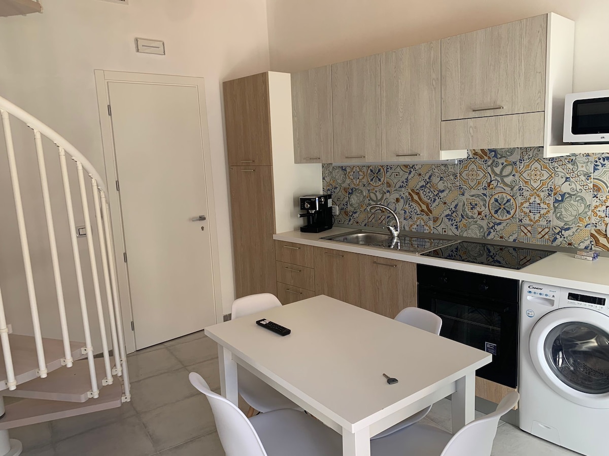 A modern kitchen area features light wood cabinetry and a decorative tile backsplash in warm colors. A dining table with four chairs is positioned in the center. Essential appliances, including a microwave and a washing machine, are visible, with a coffee machine on the counter.